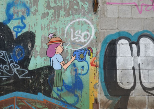 graffiti on the back of the Tower Automotive building, a tall brick building - a woman with purple hair wearing a brown and red hat.  She is holding a blue watering can.  She has an anchor tattoo on her upper arm. 