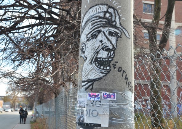 On a concrete telephone pole, a black and white drawing of a man's head.  The word Others is coming out of his mouth. 