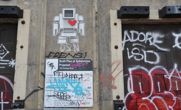 wall of a building with a wheatpaste lovebot and a standard city of Toronto development proposal sign.  Some tags are also on the building. 