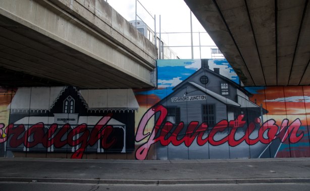 part of mural under subway bridge, large red cursive letters that say Scarborough Junction.  A picture of an old Scarborough post office as well as a wood building that was a general store. 