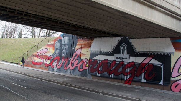 a woman is walking past part of mural under subway bridge, large red cursive letters that say Scarborough Junction.  A picture of an old Scarborough post office and two very much larger than life people (man and woman) looking east. 