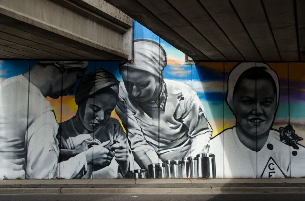 mural under subway bridge, showing woman munitions workers from the era of world war 2.  They are wearing white tops and white hair coverings. 