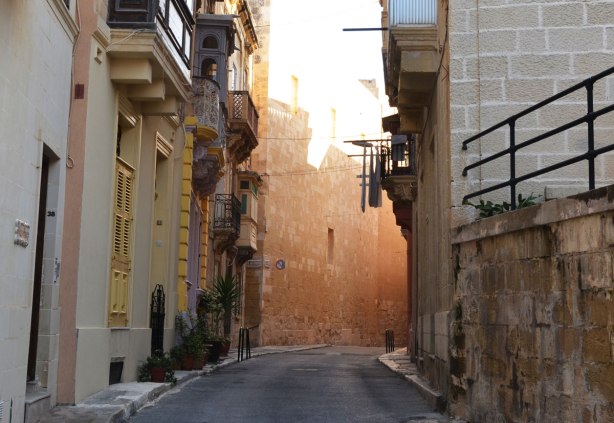 The sun shines from the end of an alley, limestone walls, laundry hanging from the balconies