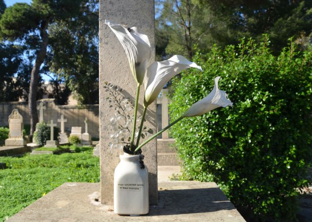 tombstone in a cemetery, with a vase of callia lilies in front of it.
