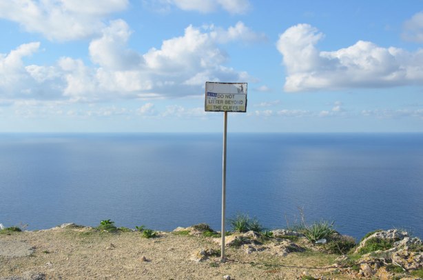 A small sign on a metal pole in the edge of a cliff. The Mediterranean Sea is behind, blue sky. The sign says Do Not Litter Beyond the Cliffs.
