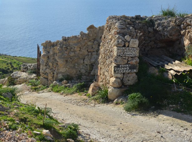 A narrow bumpy dirt road with a stone wall beside it. On the wall are a number of handpainted signs on wood that say private in a number of languages.
