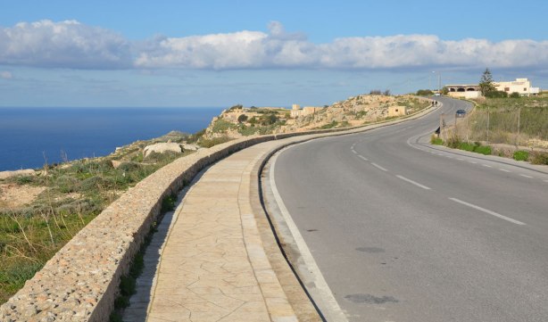A two lane paved road that runs along the top of a small cliff at the edge of the sea.