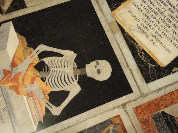 black, white, orange, gold tiles form memorials in the floor of St. Johns Co Cathedral in Valletta Malta. There are numberous angels, skulls and skeleton motifs that cover almost every inch of the cathedral floor. A skeleton is sitting at a desk and reading a book