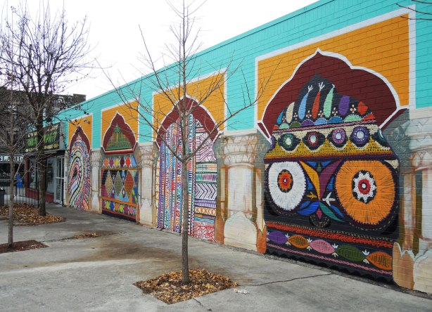 mural on the side of a one storey building showing 4 brightly coloured panels. Turquoise in the background. Each panel shows an archway between pillars. Each of the 4 has a brightly coloured pattern