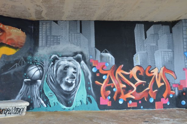 Graffiti picture of a basketball on a tree stump with a bear beside it. Large skyscrapers are in the background. A red and orange tag is also in the picture.