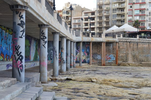 Sea wall with sidewalk above. The sidewalk is supported by concrete pillars. Rocks along the shore are in the foreground and the apartment buildings of Sliema are in the background.