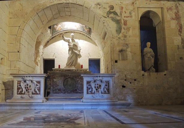Inside a grotto, A statue in a niche in the wall on the right and another statue on an altar under an arch on the left