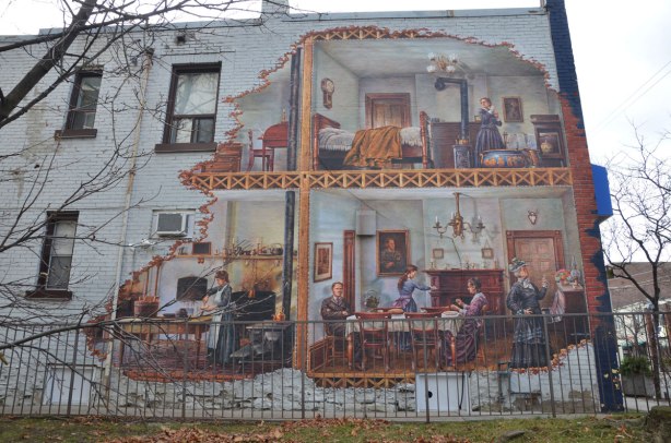 mural on the side of a two storey white brick building.  The picture looks like the outer wall has been removed to reveal a family house from the early 1900s.  A cook is working in the kitchen, 