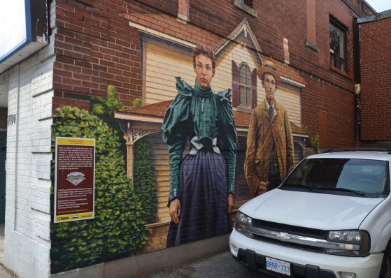 A mural of a man and a woman standing outside a two stroey farm house.  A white car is parked in front of the mural and it blocks the bottom right of the picture. 