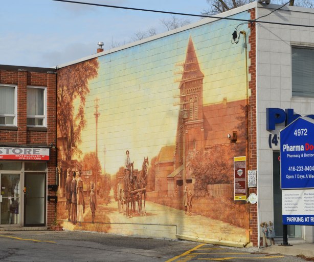 large mural on the side of a building that shows people in old fashioned clothes walking down a street.  A man in a horse drawn wagon is coming down the street. 