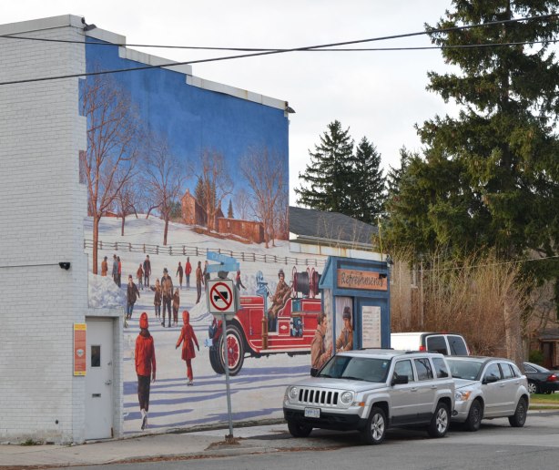 looking across the street at a mural on the side of white brick building, a winter scene, some people are skating, lots of bright red jackets, there is also an old fashioned fire engine with firefighters sitting in it.  At the right edge of the picture is a small wood hut with a sign that says Refreshments on it. 