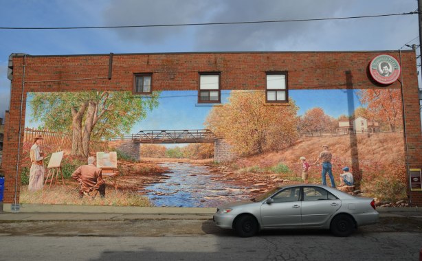 A large mural of a creek.  On the left back are two painters with their easels set up beside the river.  On the right bank are two boys and a man