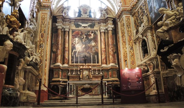 A chapel in a cathedral. An altar with six very tall candles in front of a large painting of a man on a white horse. The rest of the chapel is very ornately decorated.
