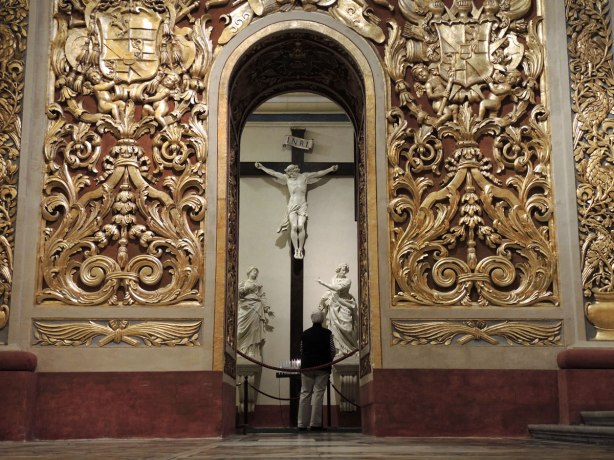 Looking across a large room with ornate gold walls towards an archway that leads to another room. In the room beyond there is a large marble crucifix with marble statues on either side of it. A person is standing looking at the crucifix with their back to the camera