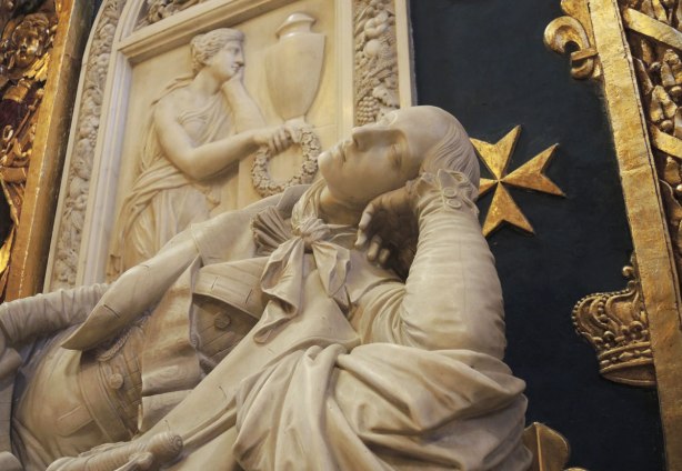 A white reclining marble male figure with his head resting on his elbow is in the foreground. A relief sculpture of a woman holding a wreath is behind him as is a blue wall with gold Maltese crosses on it.