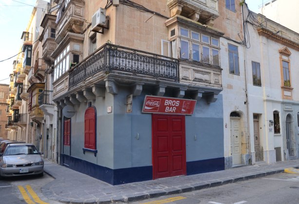 A building on the corner of an intersection. It is light blue with a dark blue band around the bottom near the sidewalk. The doors and two windows are covered with red shutter. A coke sign is above the door - the sign is horizontal and also has the name of the bar written in white letters, Bros Bar.