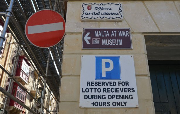 Four signs on the side of building. A red and white circular no entry road sign, a street name sign, a sign pointing towards the Malta at War Museum, and a reserved parking sign on which there is a spelling mistake.