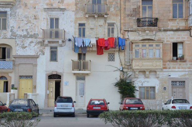 Laundry hanging from a balcony in Valletta Malta, limestone buildings and painted shutters. Four small cars parked beside the building.