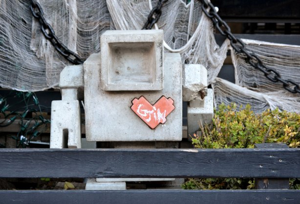 small concrete lovebot standing in a small patch of garden behind a low black wood fence.
