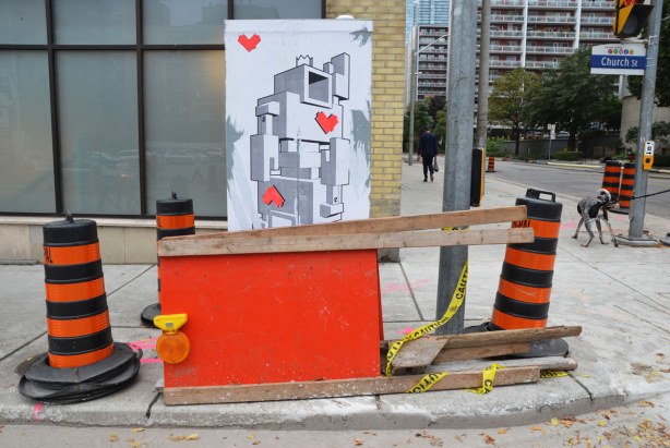 lovebot playing card on a wall. In front of it are some orange and black traffic cones along with an orange construction sign on its side.