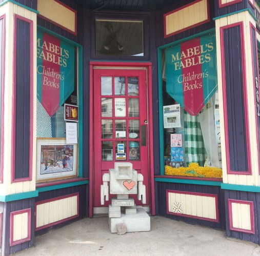 A small concrete lovebot stands in the doorway of a childrens book store called Mabels Fables.