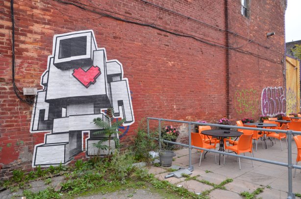 A very large lovebot on a brick wall. He looks like he is walking away from a restaurant patio with its black round tables and bright orange chairs.