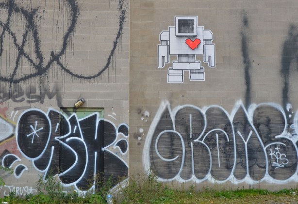 Wheatpaste lovebot up high on a concrete wall. There are some black and white tags below him.