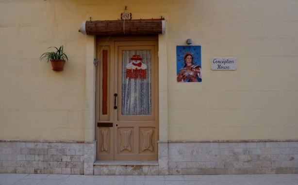 Doorway, wood door on pale yellow house. plant on wall on one side of the door, a picture of Mary on the right. A Santa decoration is on the door.