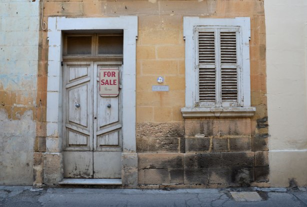 House for sale. An old grey door and old grey shutters on a wondow. For Sale sign on the door.