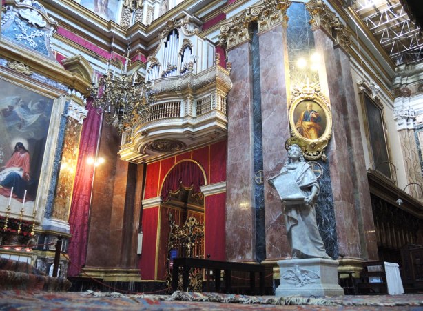 interior of the cathedral in Mdina, statues and stone work. Parts of the walls are covered in red fabric.