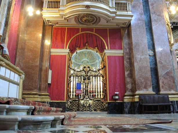 Interior of the cathedral, carpet on a few stairs, a metal gate in front of a side chapel, some walls covered in red fabric, marble work,