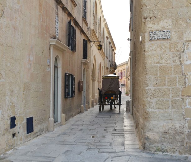 horse drawn carriage passes through a narrow lane, yellow limestone buildings on either side.