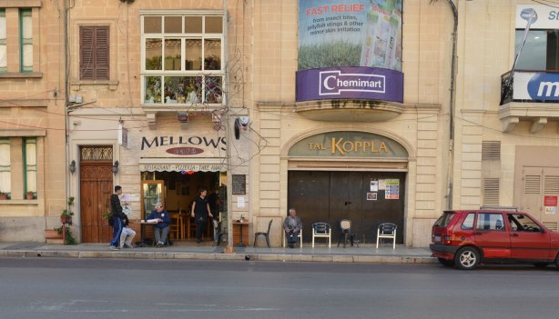 looking across the street to Mellows, a restaurant and pizzeria. There are people sitting at tables outside on the sidewalk. Beside Mellows is a store called Tal-Koppla but it is closed up.