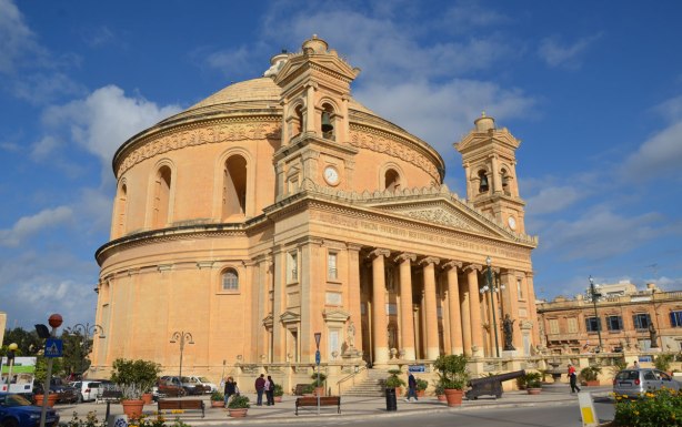 The front of Mosta Rotunda from Pjazza Rotunda, the front of the church with its tall columns can be seen. The round lower part of the dome is also visible.