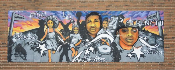 A large mural on a red brick building, painted to celebrate the United Nations Universal Declaration of HUman Rights. This one includes the words strength, love, respect, unity. It shows a 5 people engaged in various activities.