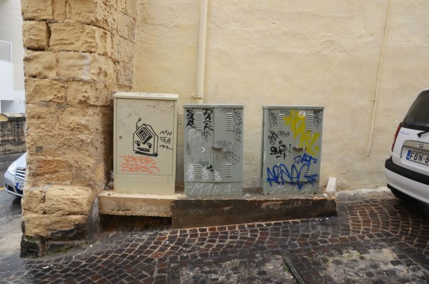 Three electrical boxes in a row, beside a cobblestone street. They have graffiti on them.