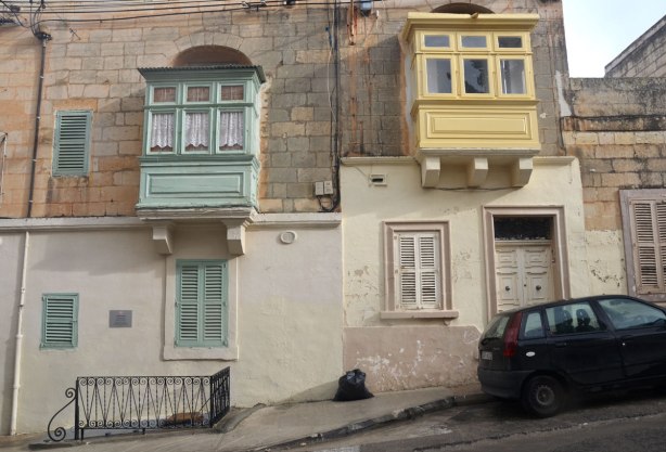 looking across the street to two houses, each with a wooden balcony structure overlooking the street. One is pale green and the other is yellow.