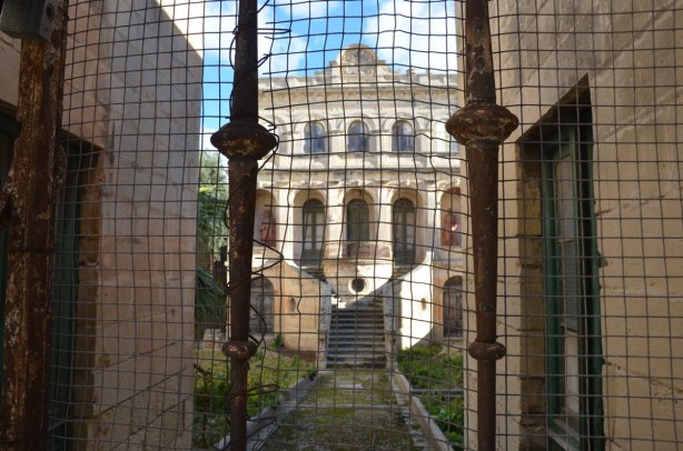 an old, large, ornate, 3 storey abandoned stone house in Rabat, behind an old rusty wire mesh gate.