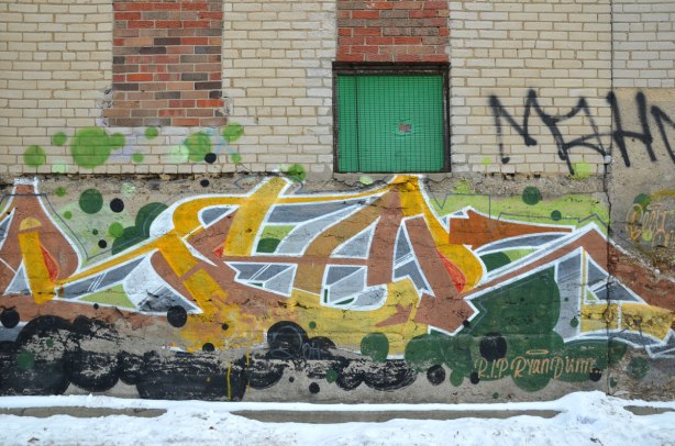 Old graffiti taf in yellows and blues on a grey brick wall.  Two windows have been bricked over in red brick.  One window has a green wood covering.  It includes the words "RIP Ryan Dunn"