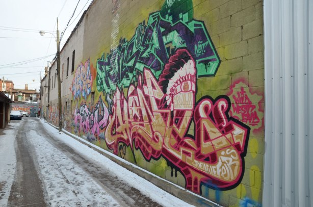 looking down an alley with the side of a two storey building being the dominant part of the picture.  It has a large number of colourful graffiti tags painted on it. 
