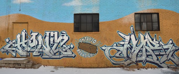 street art on a wall.  In the middle is a brown loaf of bread with the words Ontario Bread Co written around it.  There is a black and white tag on either side.  two windows and a door of the building are also in the photo. 