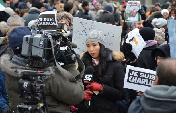 A large TV camera is filming a reporter from CP24.  People behind the reporter are holding Je Suis Charlie signs. 