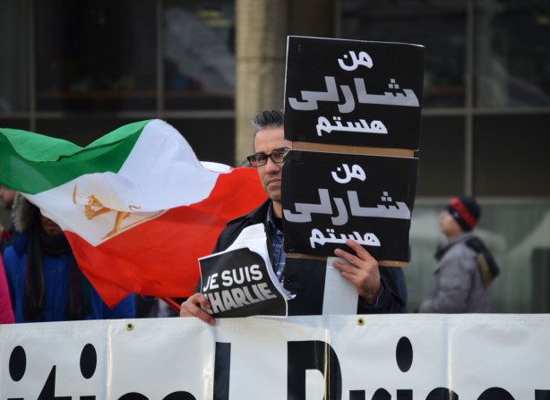A man is holding three signs.  One says 'Je Suis Charlie' and two are in arabic.  He standing behind a large white sign withblack letters.  The words can't be read in this photo.  An Iranian flag is being held by someone standing behind him. 