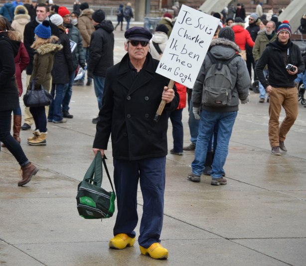 A man is wearing yellow clogs and holding a sign that says Je Suis CHarlie. Ik ben Theo Van Gogh