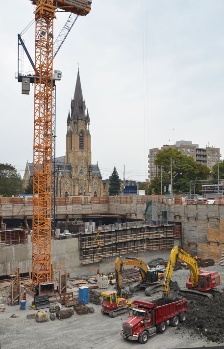 A construction site, hole in the ground, large yellow crane, red dump trunk and a large fence around the site.  A church is in the background. 